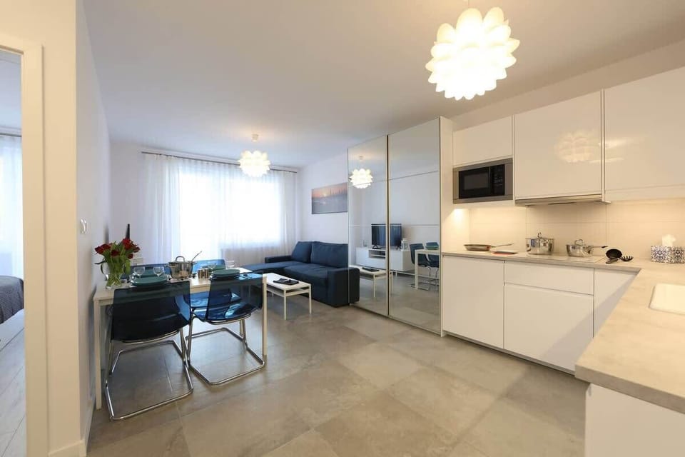 An open-plan living room with a gray sofa, a dining area, and a white modern kitchen in the background.