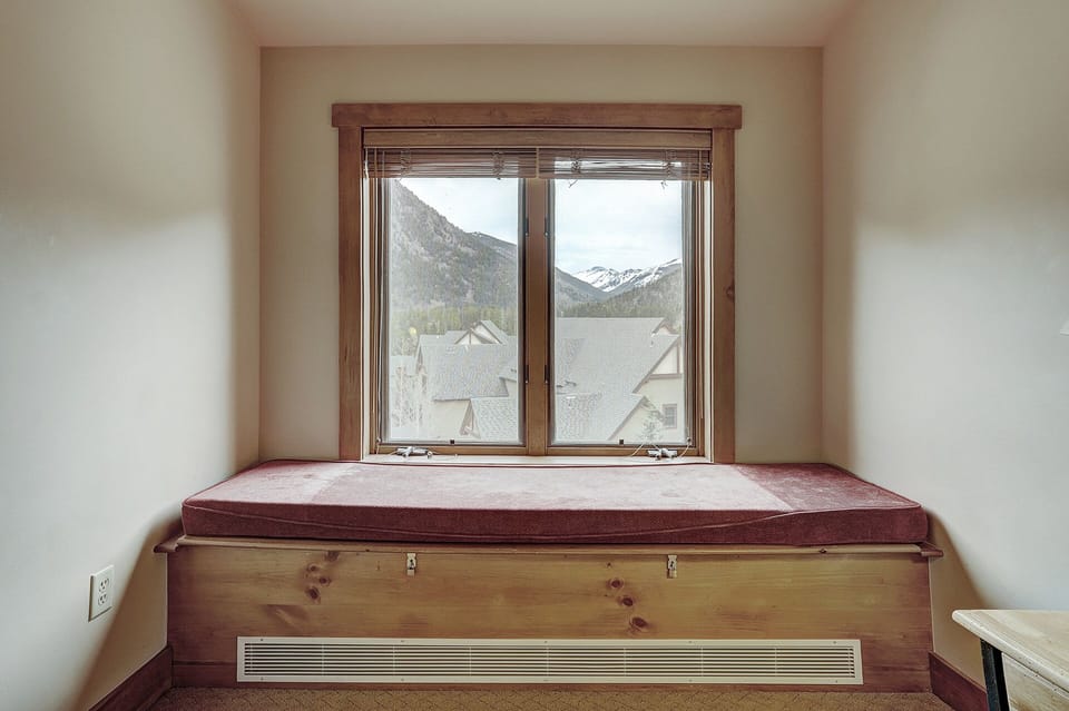 A small room with a window seat under a double-pane window. The window has a wooden frame and overlooks rooftops and snow-capped mountains in the background. The window seat has a maroon cushion.