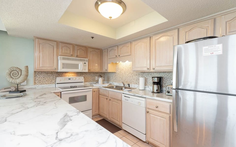 This kitchen features light wood cabinetry, white and stainless appliances, a coffee maker, and elegant countertops