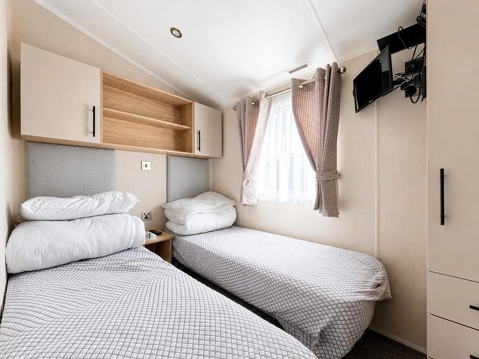 Two single beds with patterned gray bedding sit beneath wood and cream overhead cabinets in the bright bedroom.