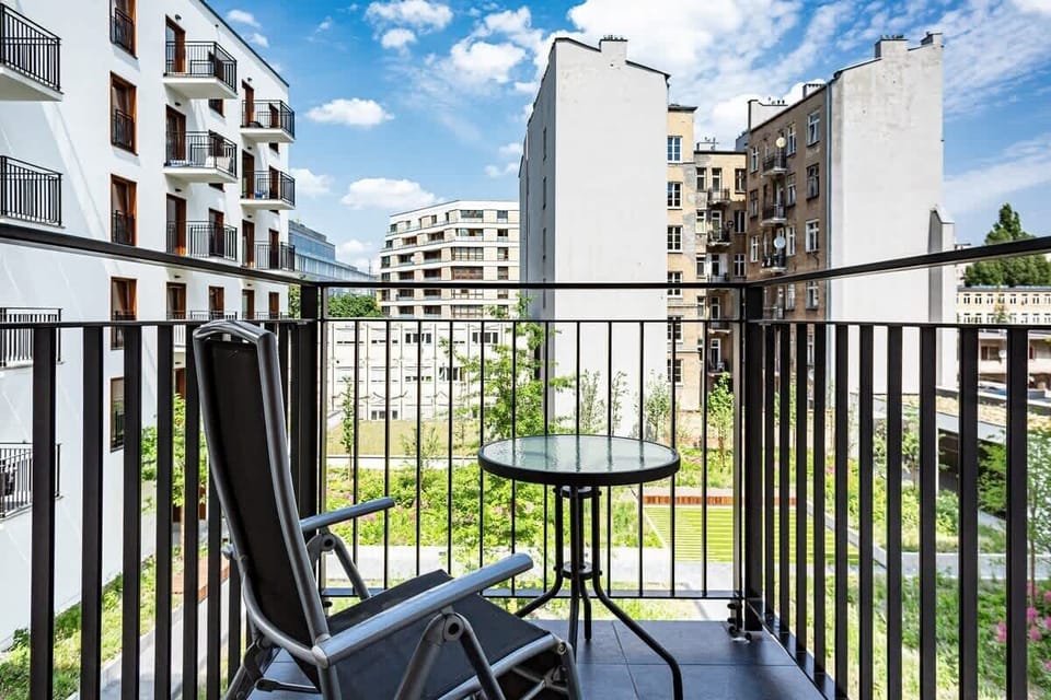 The image shows a residential balcony with outdoor furniture. In the background, there's a modern apartment complex surrounded by greenery and open spaces.