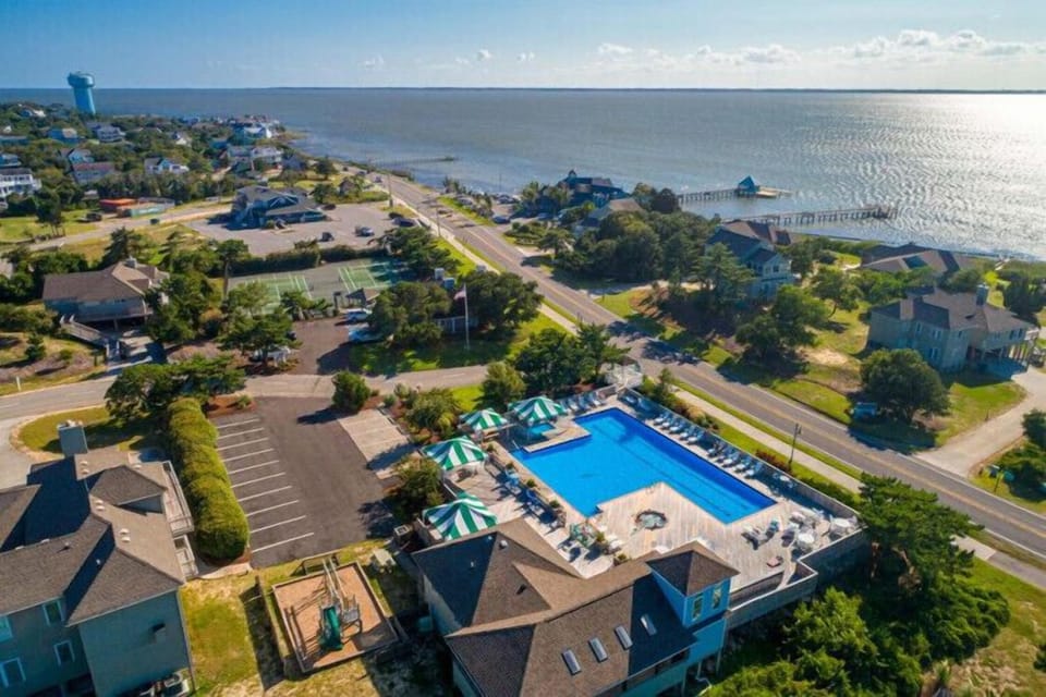 Aerial view of waterfront property with pool, located steps from the bay and beach. Sparkling blue water stretches to the horizon under clear skies.