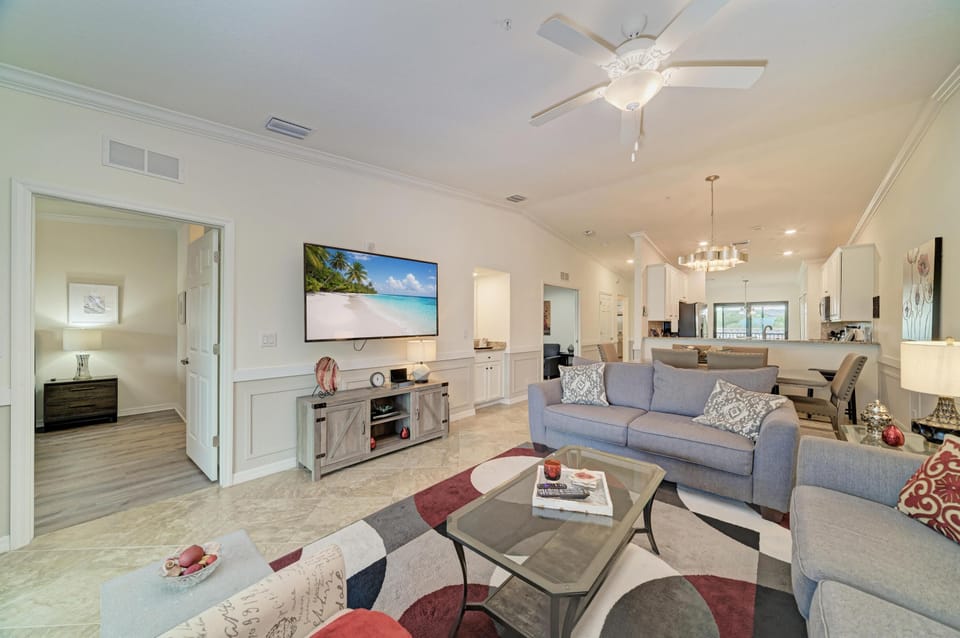 Living room with light gray sofas, glass-top coffee table, patterned area rug, and wall-mounted flat-screen TV.