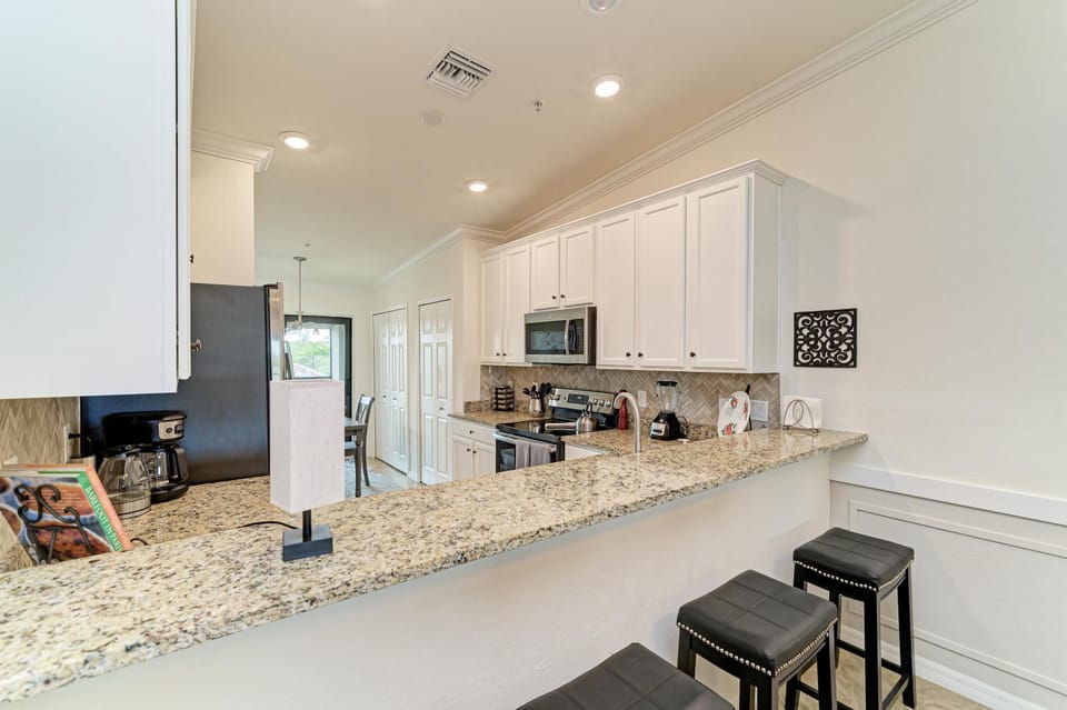 Granite countertop breakfast bar with black leather stools overlooks white cabinetry and stainless steel appliances.