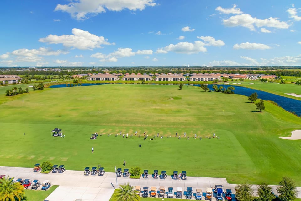 Wide outdoor view of a golf driving range with lined golfers, golf carts parked on concrete, and a blue water hazard.