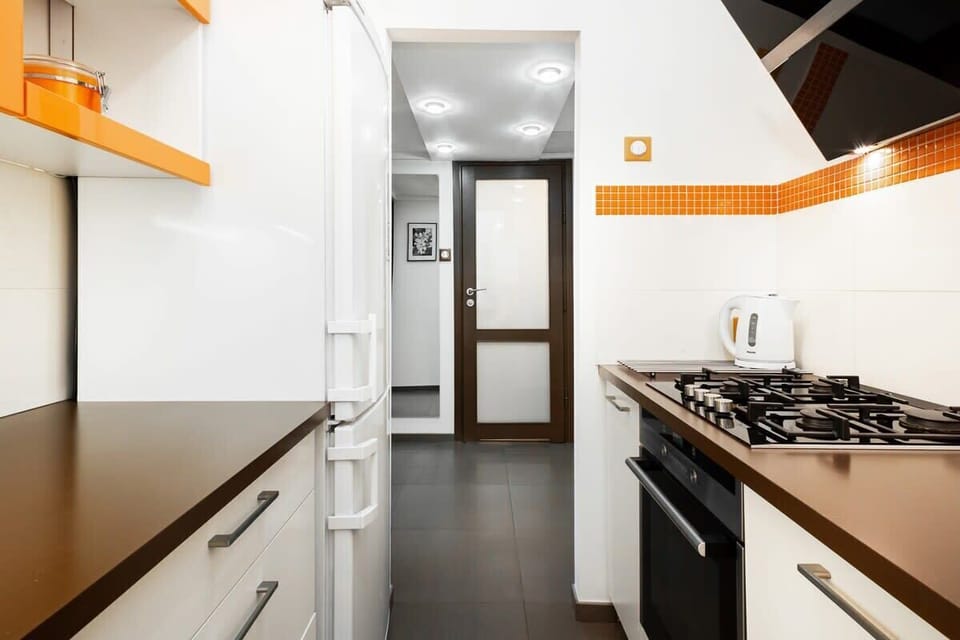 A narrow kitchen layout with white cabinets, a countertop, and an orange accent strip. The design is compact, with modern appliances.
