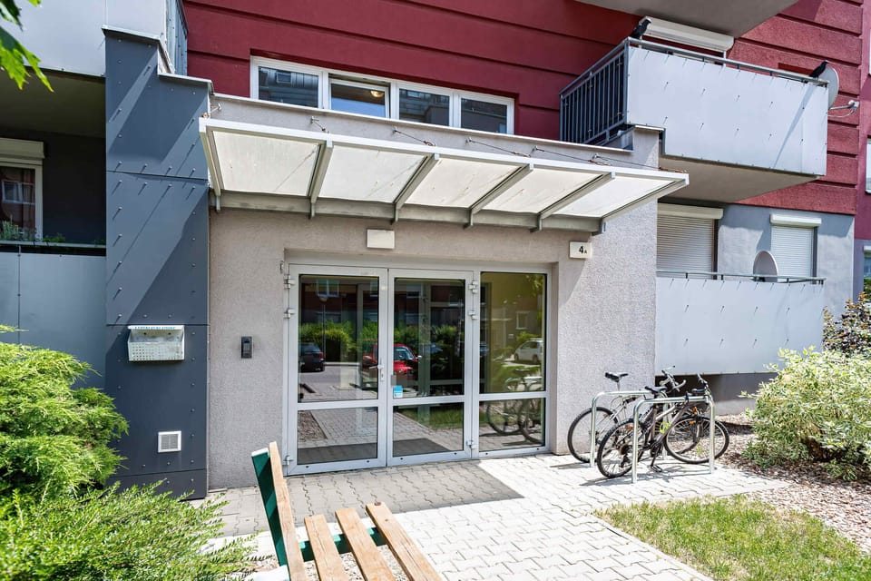 The entrance of a modern apartment building with large glass doors, a bicycle rack, and well-maintained greenery framing the space.