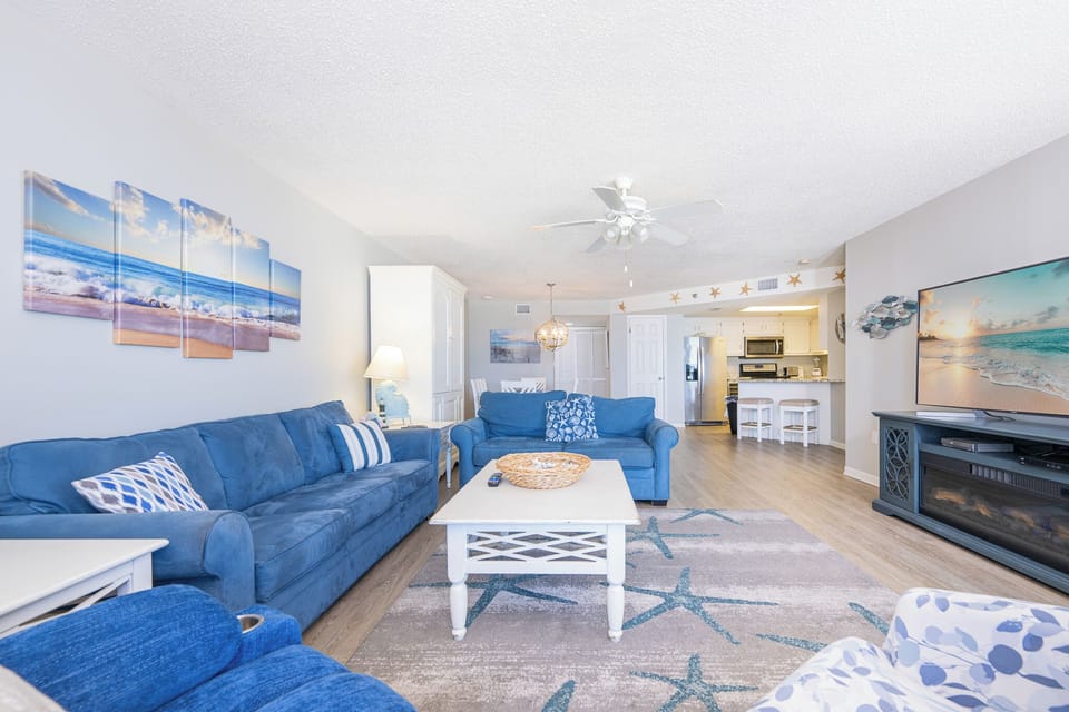 The living room features cozy blue sofas, a light-filled atmosphere, and a serene beach view through the sliding glass door