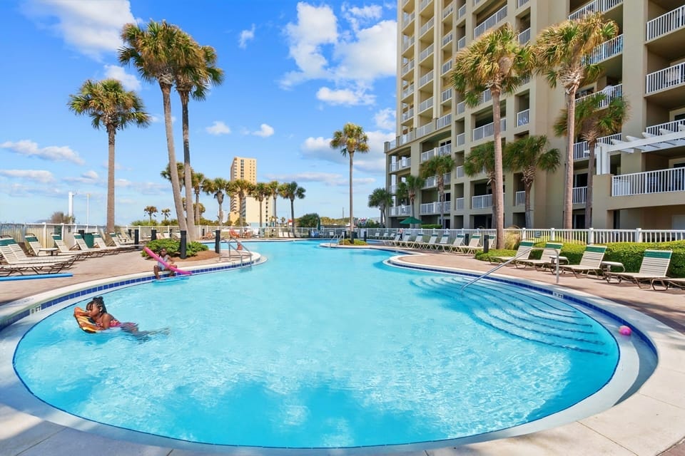 Expansive resort-style pool winds past swaying palms and private balconies, with sparkling water and sunny lounging areas that invite you to relax in tropical comfort.