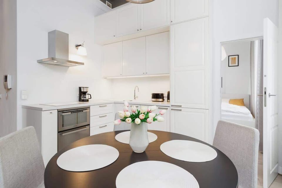 A compact dining area with a round table and chairs. The kitchen can be seen in the background, with white cabinetry and a clean layout.