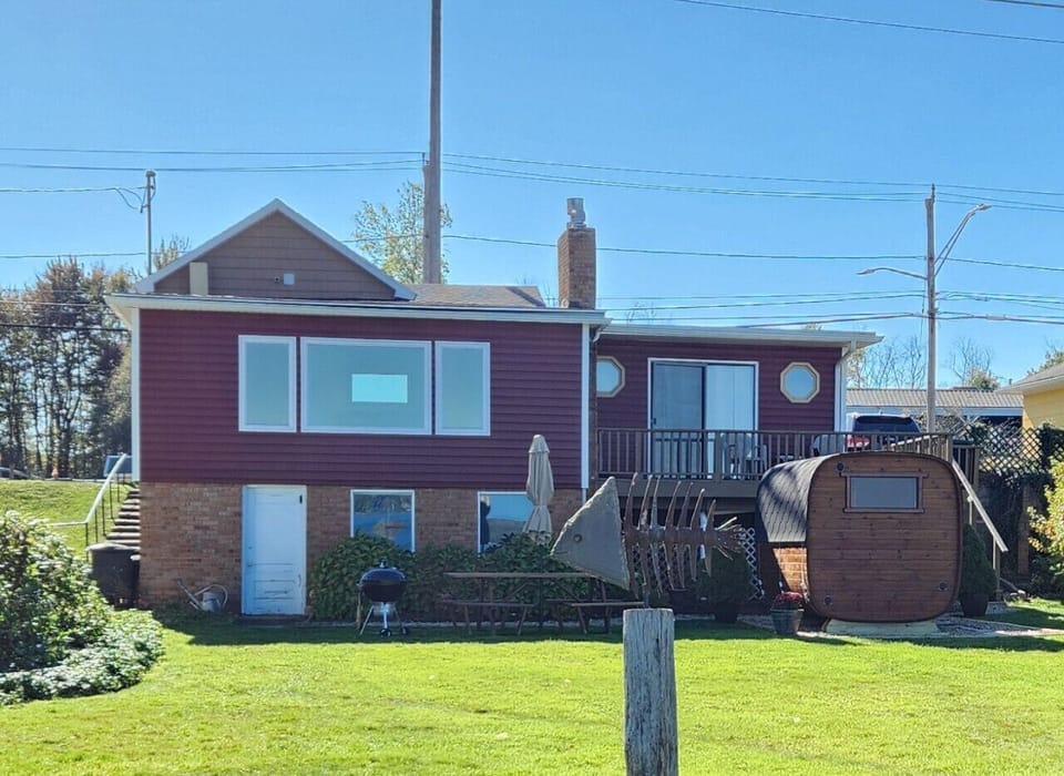 View on a red sided house with wooden cube Sauna, picnic table and a fish art work in front - blue sky
