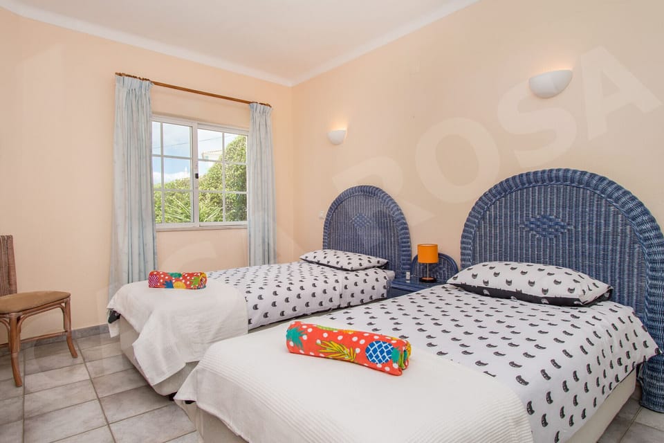 Twin bedroom at Casa Rosa villa with two single beds, colourful bedding, wicker armchair, and natural light from the window, ideal for children or guests staying near Carvoeiro in the Algarve