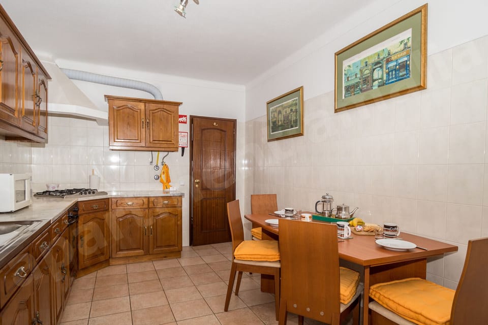 Dining room at Apartment Amalia in Carvoeiro, with a wooden dining table and chairs set for a meal, open archway through to the fully equipped kitchen, framed artwork on the walls, and tiled flooring
