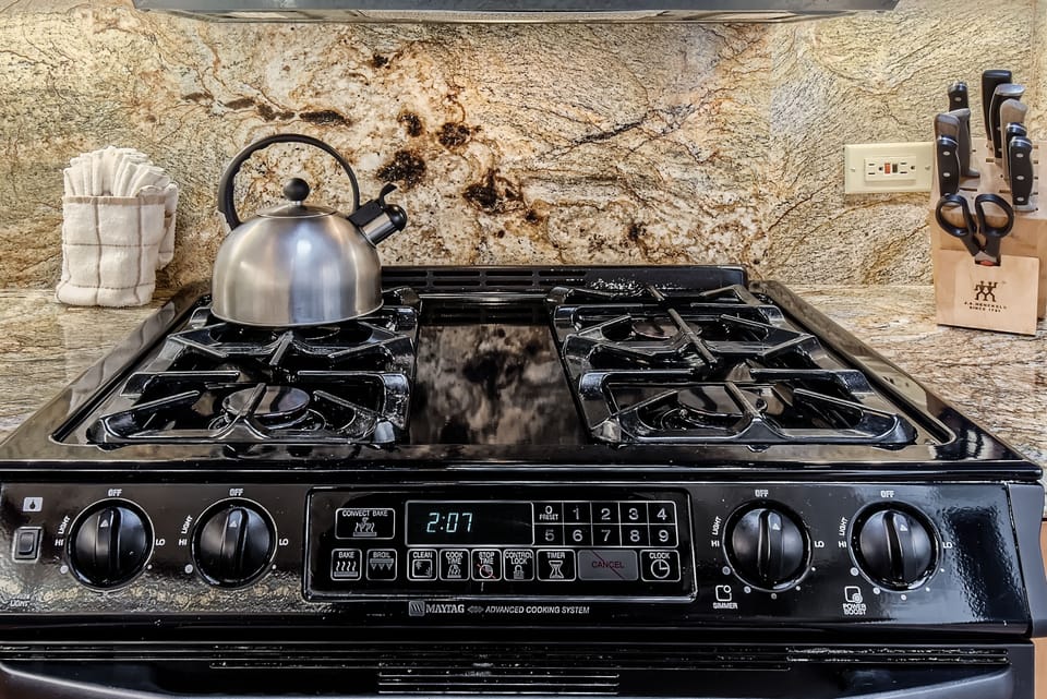 A black stove with four burners, a tea kettle, and digital controls displays the time 2:07. Knives are stored in a block on the right, and a towel holder is on the left. The backsplash features a stone pattern.