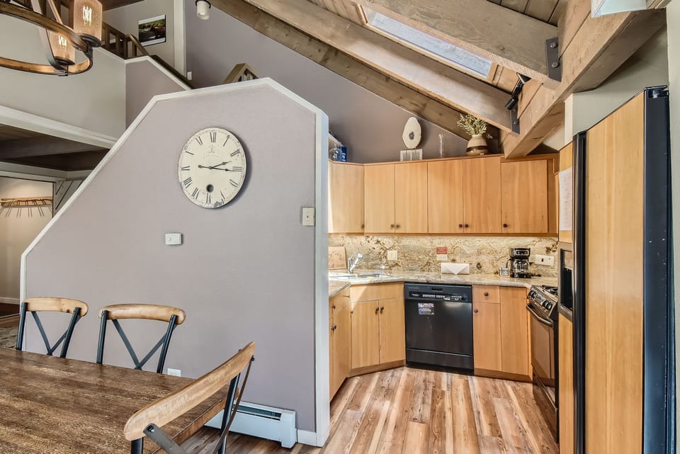 A modern kitchen with wooden cabinets, marble countertops, stainless steel appliances and a skylight above. A large wall clock decorates the adjacent wall. A dining area with a wooden table is visible.