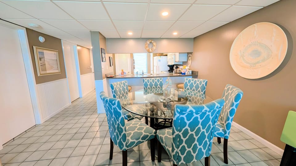 Dining area with a round glass table surrounded by six patterned blue chairs on tile flooring.