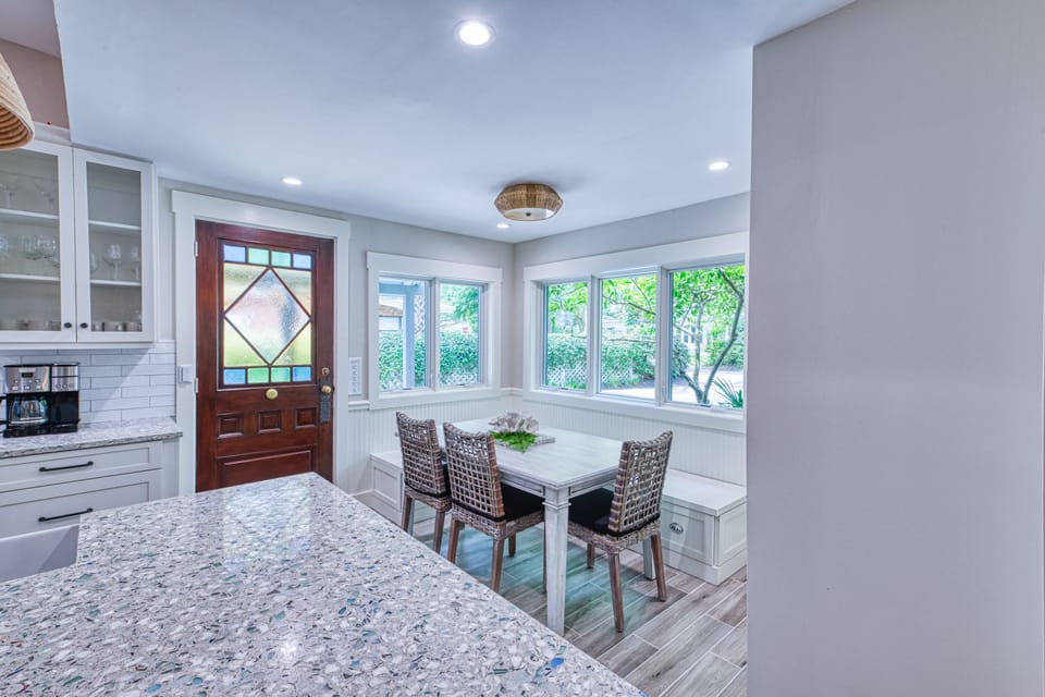 Bay Window Dining Nook and Entry View – Tabby on 12th - This corner of the kitchen captures the bay window nook, entry door, and island flow—perfect for sipping coffee, sharing beach plans, or unpacking a market haul. Bright, coastal, and made for moments.