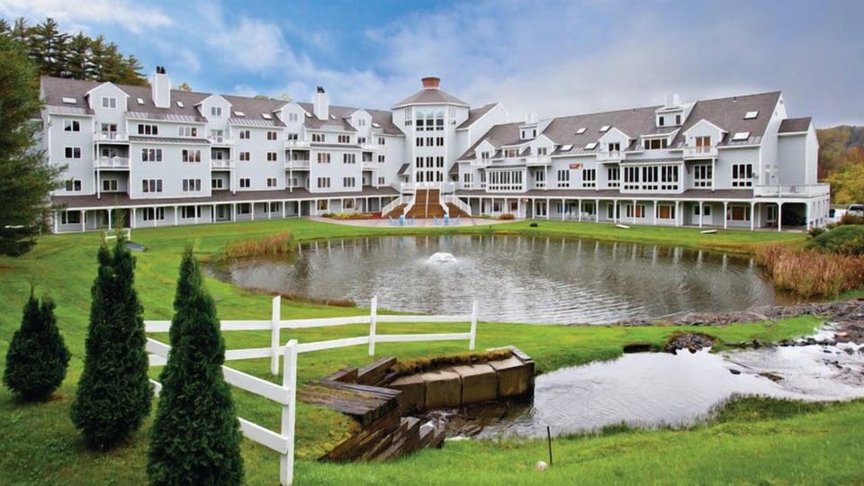 White villa building with multiple balconies and gabled roofline, set behind a green lawn and pond