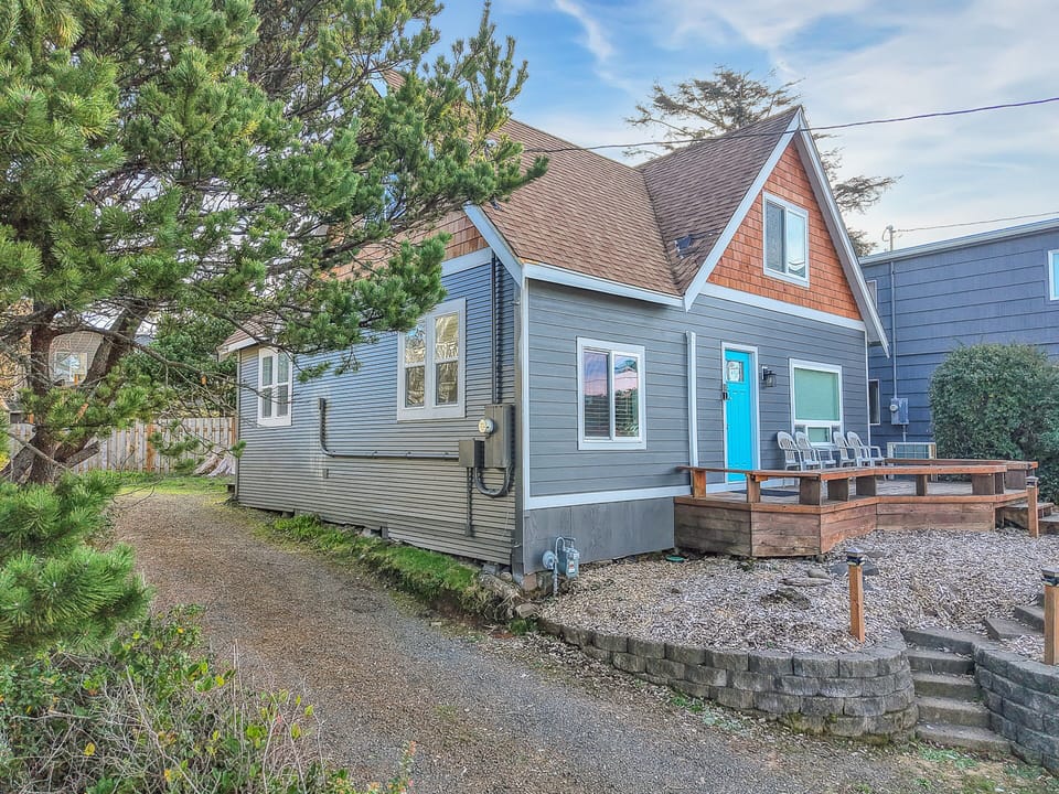 Charming coastal cottage with bright blue door and private deck, surrounded by mature trees in a quiet neighborhood setting.