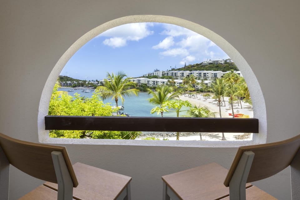 Two bar height chairs face an arched balcony opening with ocean, palm trees, and sandy beach views.