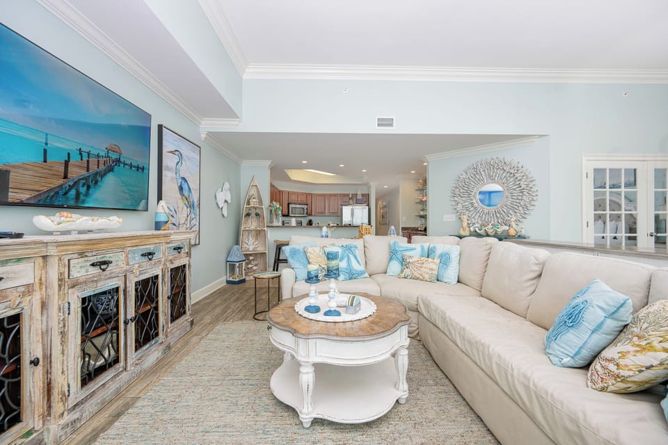 The living area features a large sectional sofa with blue and beige pillows, a flat-screen TV, and a view of the beach