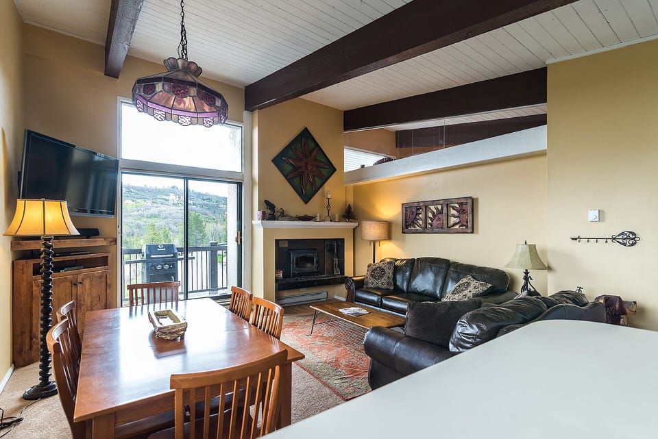 Relax in your cozy living space featuring exposed beams, a warm fireplace, and comfortable leather seating with mountain views through oversized windows.