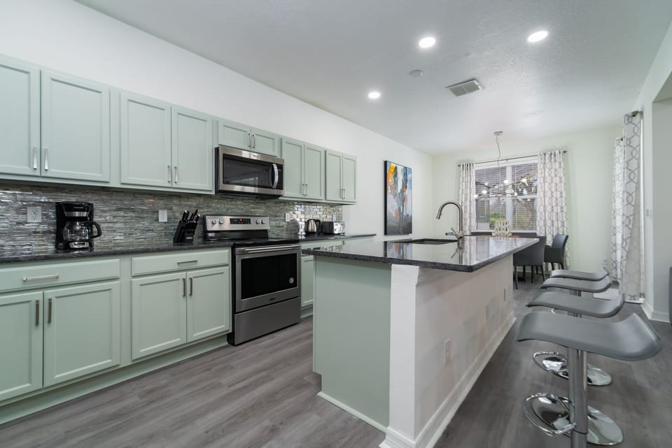 Modern open concept kitchen with mint green cabinets, stainless steel appliances, mosaic tile backsplash, and large granite island with bar seating.