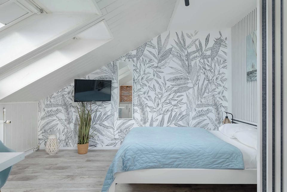 A close-up of the bed in the bedroom with blue bedding, wallpaper featuring leaf designs, and a TV mounted on the wall.