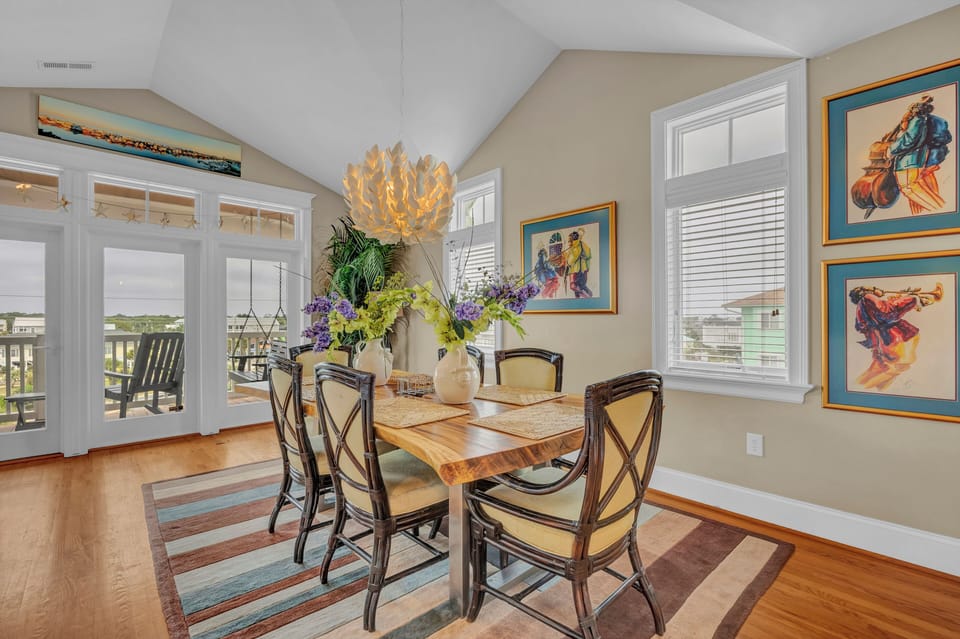 Gather around this inviting dining table under a stunning chandelier, where natural light pours through expansive windows, offering views of the outdoor living space. Enjoy your meals in style, surrounded by tasteful artwork and coastal charm.