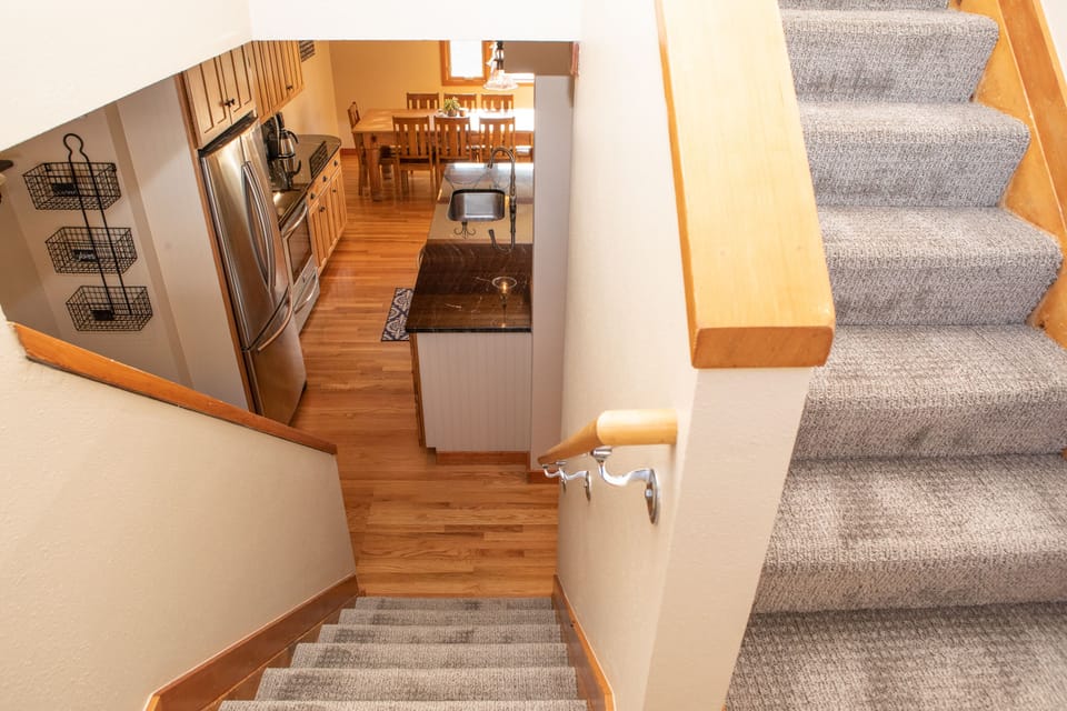 View from the top of carpeted stairs looking down into a kitchen with stainless steel appliances, wooden cabinets, granite countertops, and a dining area.