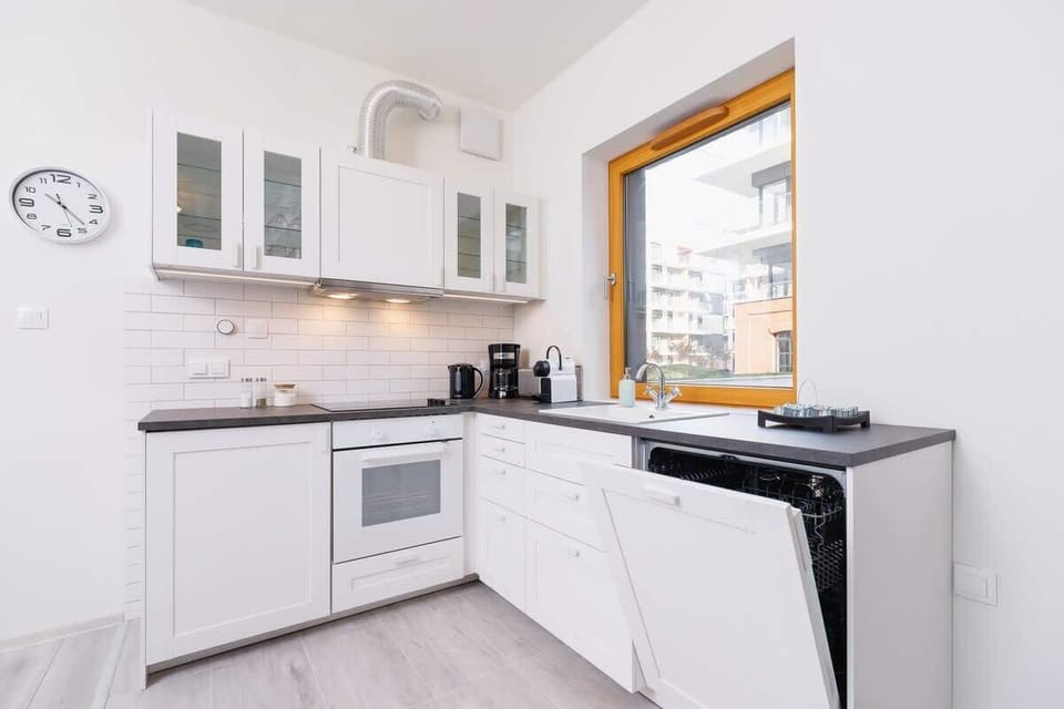 A compact kitchen with white cabinetry and modern appliances. It features a large window and minimalistic design, providing a clean and functional cooking space.