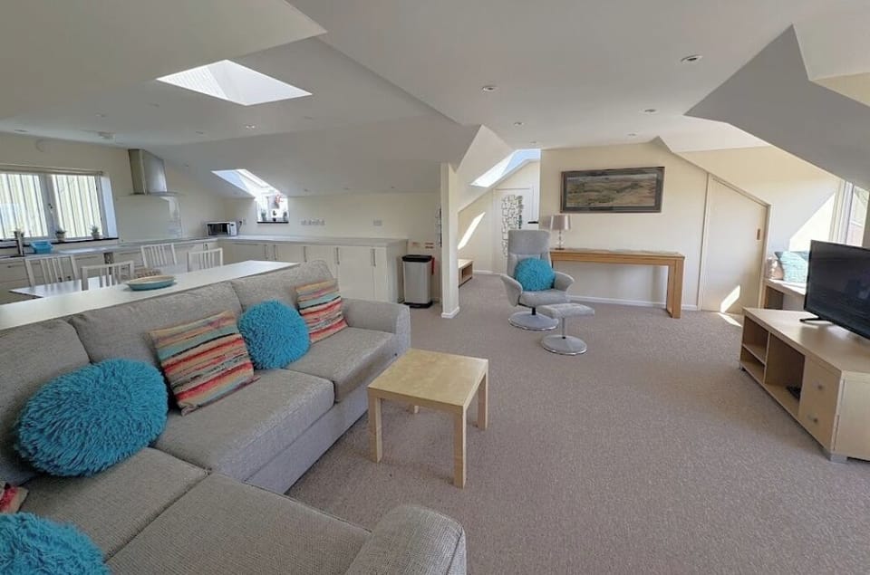 Living area showing sofa, chair, TV and some of the kitchen area