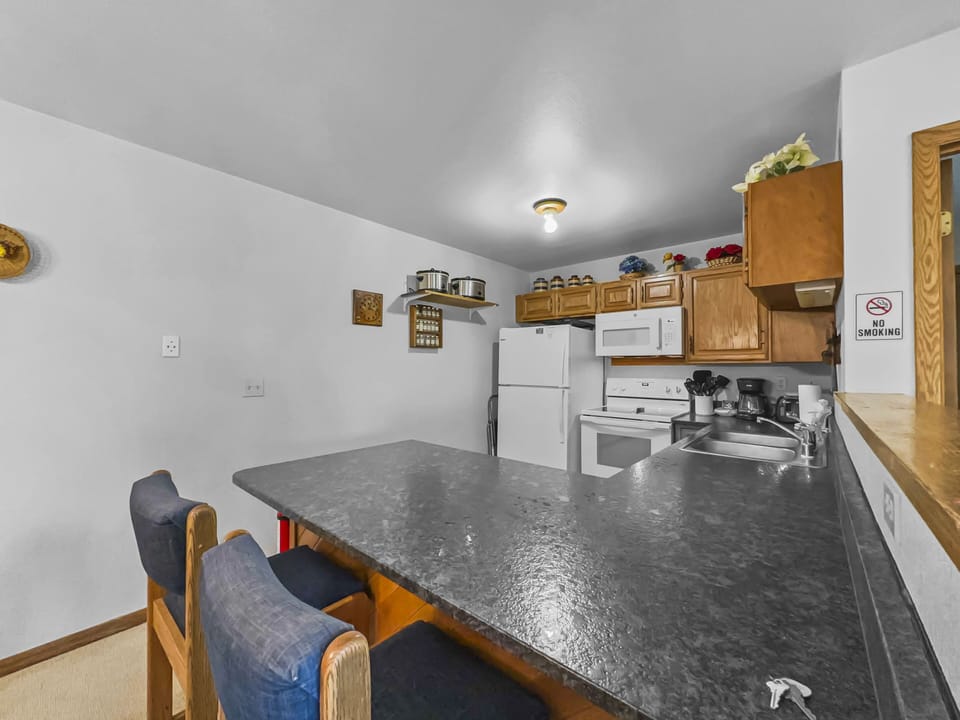 A kitchen with wooden cabinets, white appliances (refrigerator, microwave, and stove), and a gray countertop with two blue cushioned bar stools. A "No Smoking" sign is posted on the wall.