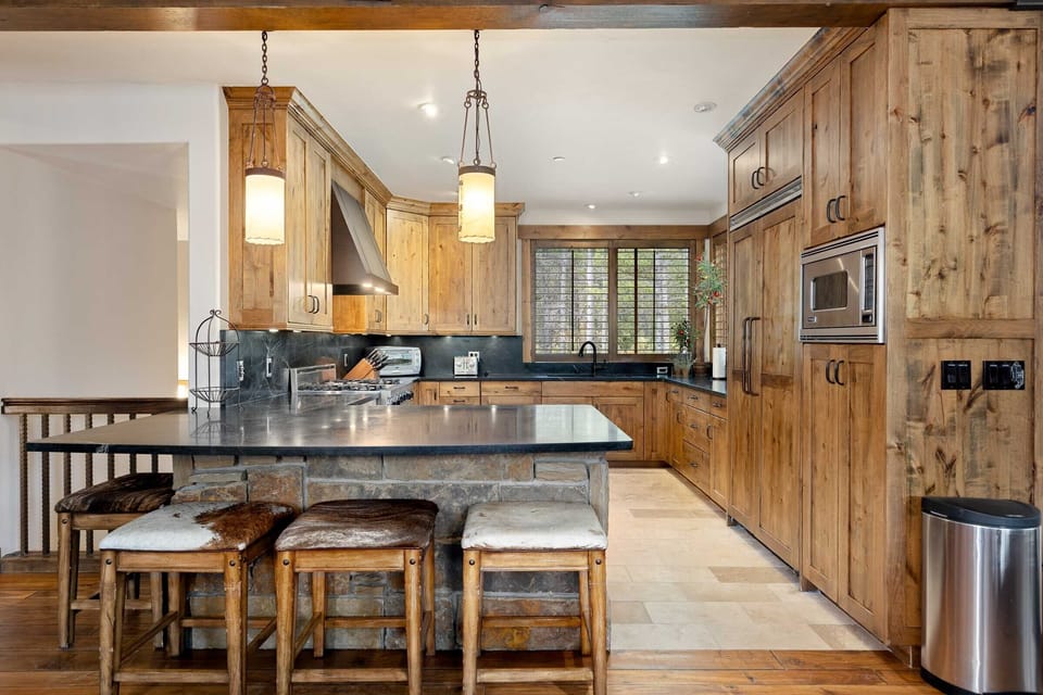 A rustic kitchen features wood cabinets, stainless steel appliances, an island with seating, and stone countertops. Natural light enters through a window by the sink.