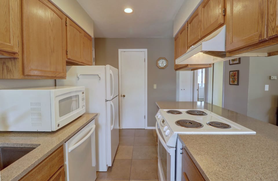 A kitchen with wooden cabinets, white appliances including a refrigerator, oven, and microwave, and a beige countertop. A wall clock and some small framed pictures are on the wall.