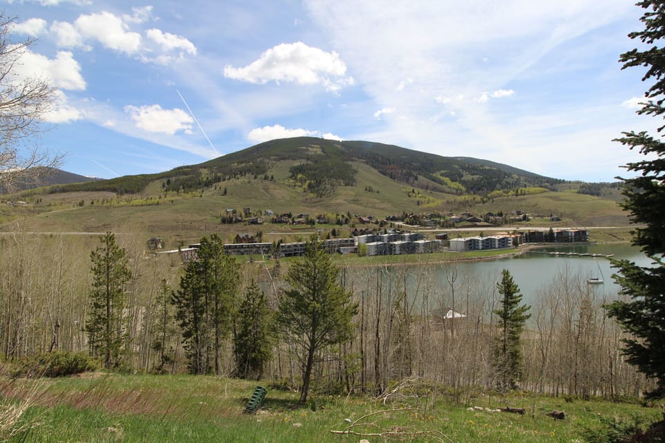 A scenic view of a small town nestled in a valley, surrounded by green hills and trees, with a lake in the foreground under a partly cloudy sky.