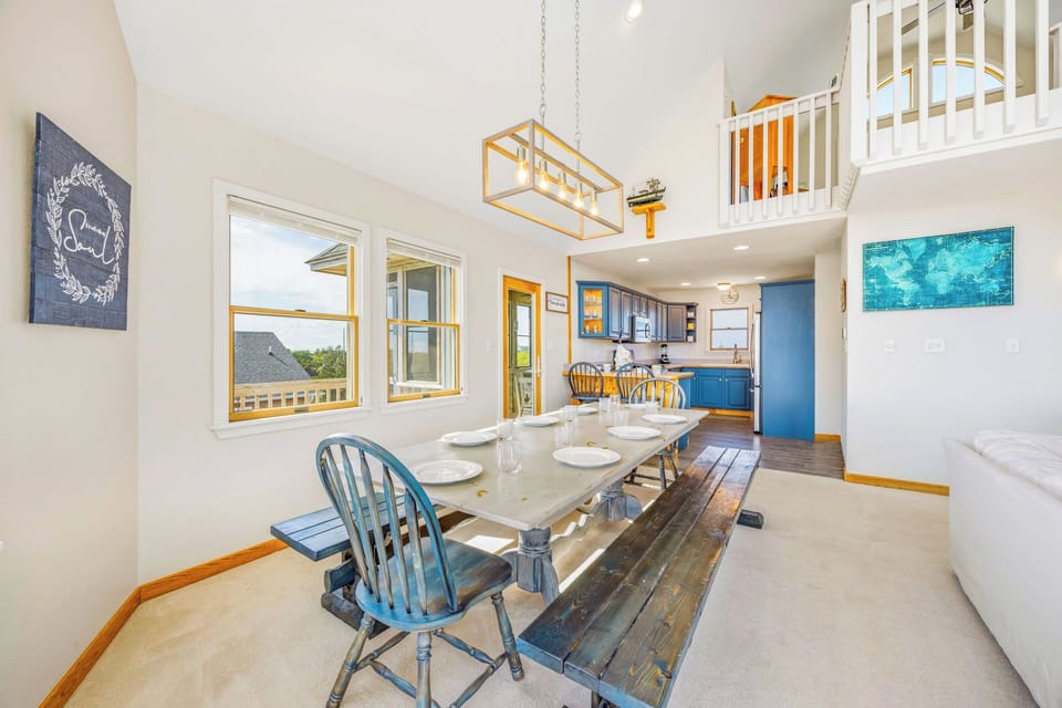 Gather around the rustic dining table where natural light pours in, highlighting cozy coastal touches and an open layout that flows into the kitchen and loft above.