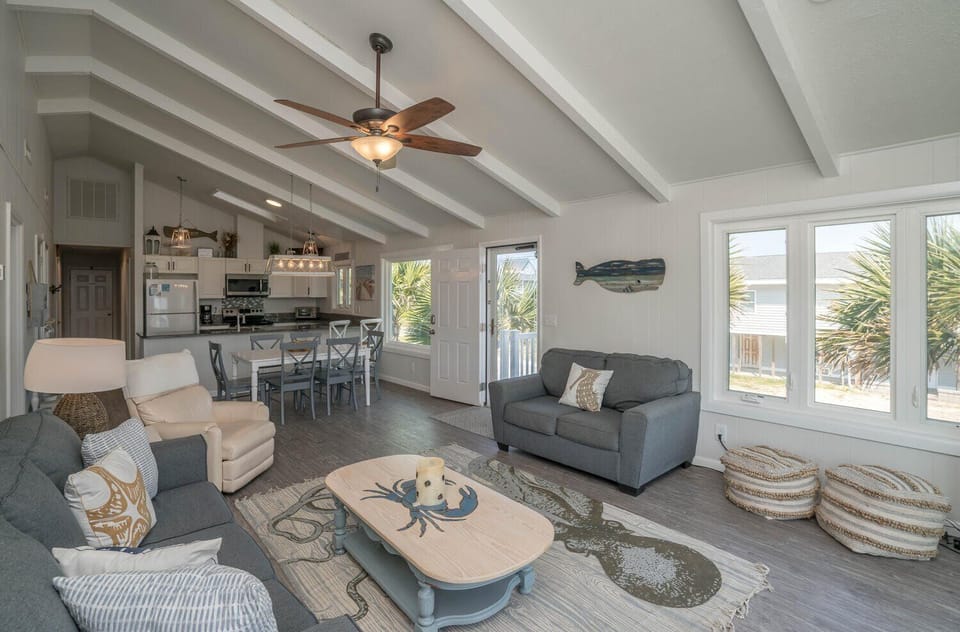 Spacious Oak Island, NC living area with vaulted beam ceilings, ceiling fan, gray sofas, recliner, coffee table, coastal rug, dining table, white kitchen, windows, beach décor, and door opening to a sunny deck.