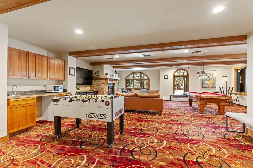 An expansive game room invites you to unwind with foosball, pool, and cozy lounge seating, all warmed by rustic beams and arched windows overlooking the outdoors.