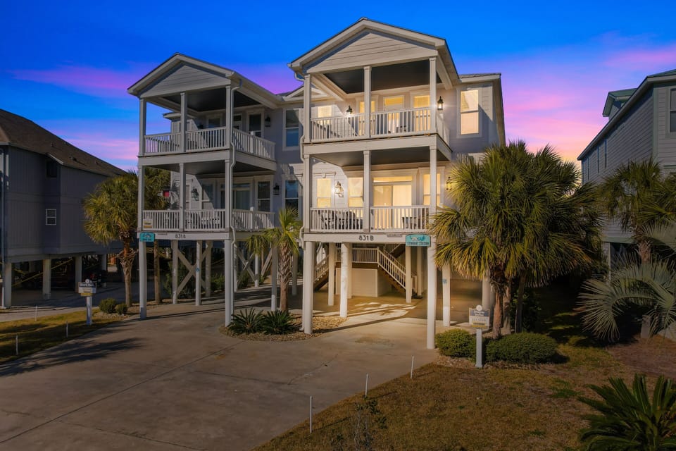 Charming coastal beach house with elevated design, multiple balconies, and tropical palm landscaping at twilight.