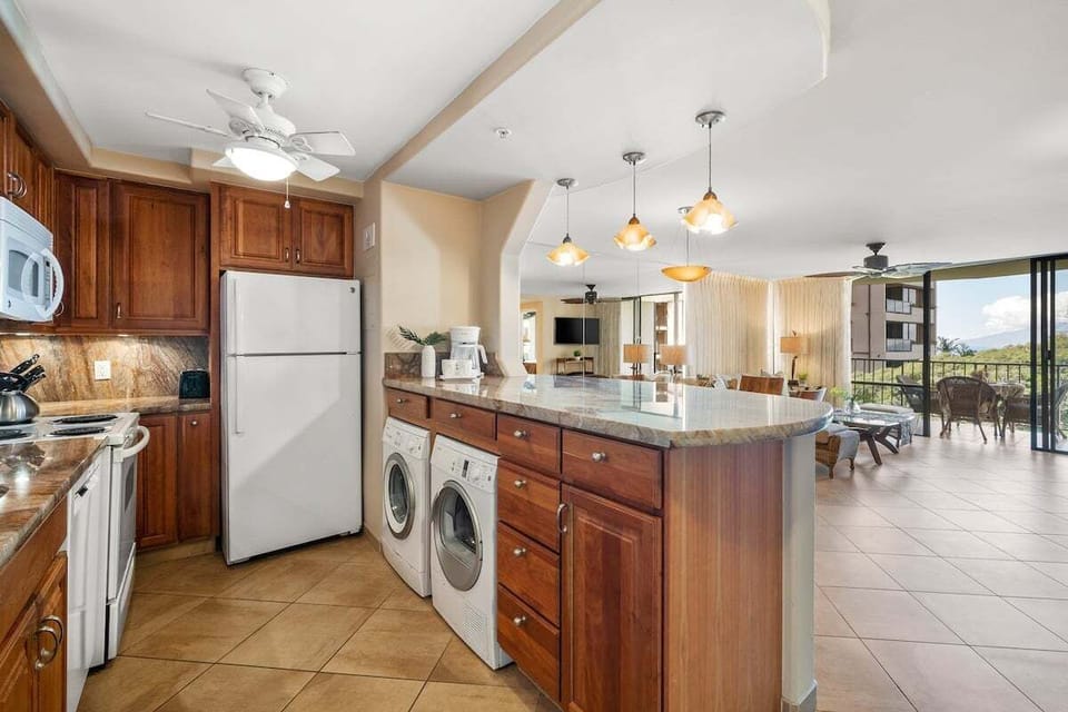 Kitchen with refrigerator, washer, dryer, granite countertops, and pendant lights, open layout connecting seamlessly to the dining and living areas with balcony access.