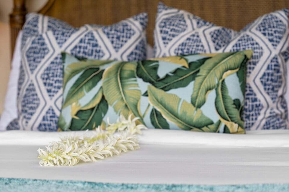 Close‑up of a bed with decorative pillows in blue and white geometric patterns and a green leaf design, a white flower lei adding a tropical touch