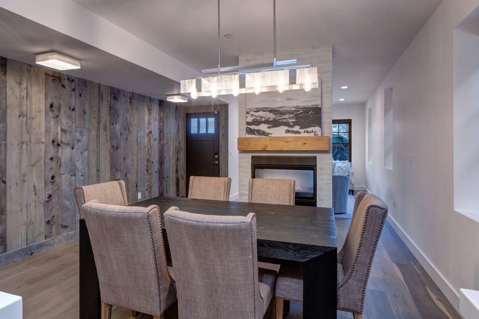 Gather for a memorable meal in our stylish dining area, featuring a large table, upholstered seating, a modern chandelier, and a cozy fireplace with a wood mantel.
