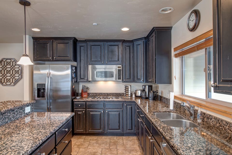 A modern kitchen with dark wood cabinetry, granite countertops, stainless steel appliances, and a double sink. A clock hangs on the wall above a window. A pendant light fixture is visible.