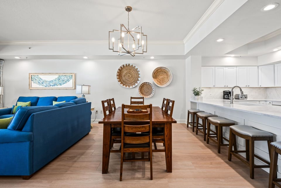 The open-concept living space flows seamlessly from plush blue sofas to a warm wooden dining table and sleek white kitchen, perfect for gathering.