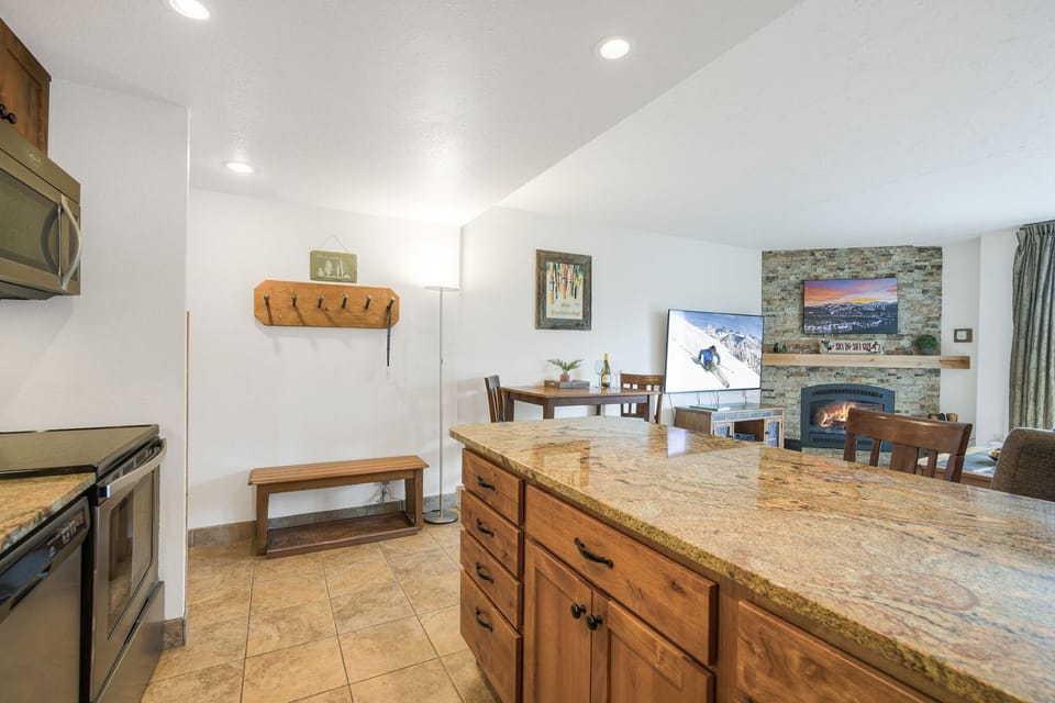 Granite kitchen island with wood cabinetry overlooks a corner dining table and stone fireplace with mounted TV.