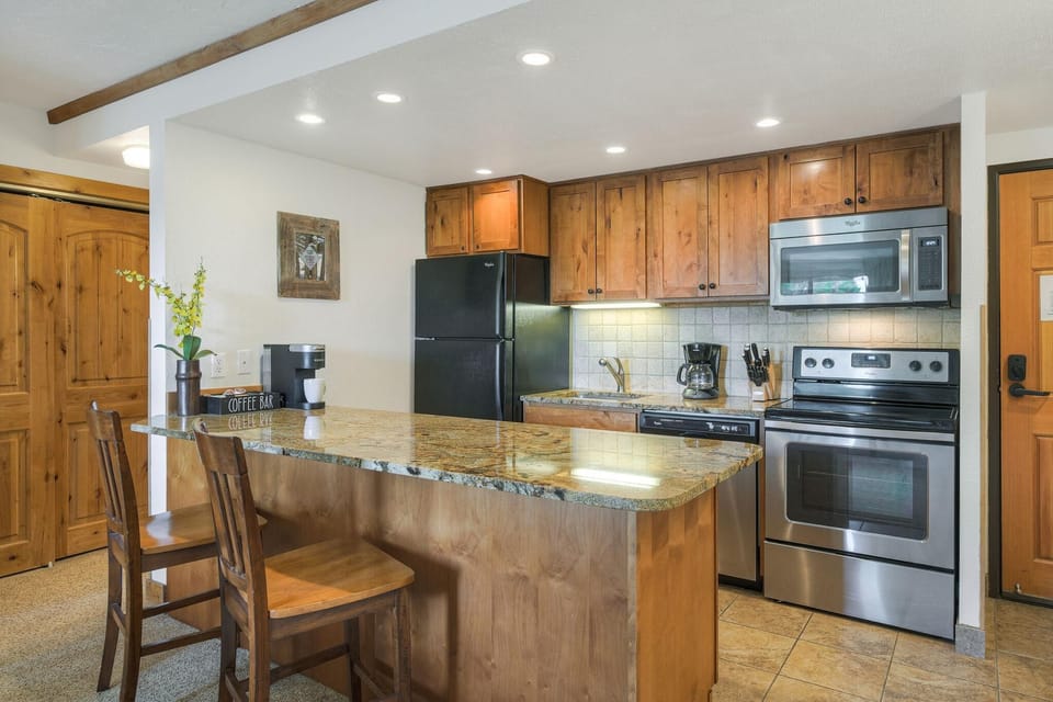 Granite countertop extends into breakfast bar with wooden stools, surrounded by warm wood cabinetry and stainless appliances.