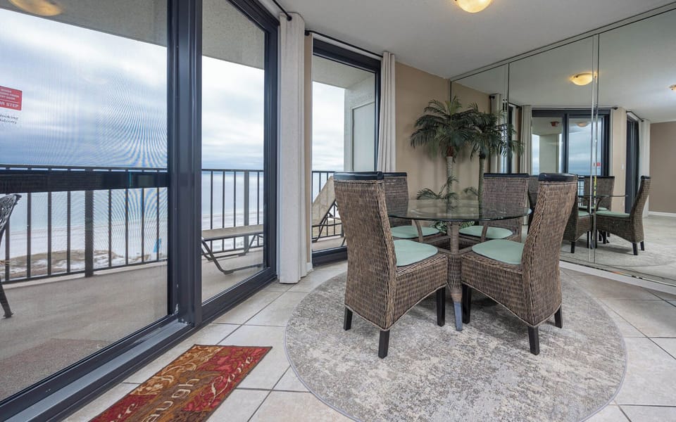 Dining area features a glass table surrounded by woven chairs, with a view of the balcony and the beach beyond