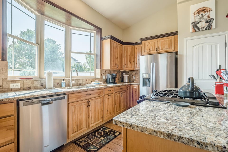 Granite countertops and natural wood cabinets surround stainless steel appliances beneath a large, sunlit window in the kitchen.