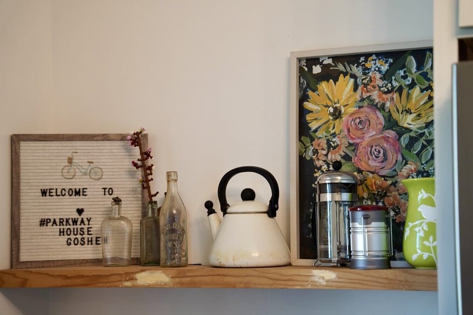 Charming shelf styling with a welcome letterboard, antique glass bottles, and a vintage tea kettle. A colorful floral painting and French press add warmth, celebrating the little historic details that make Parkway House Goshen feel like home.
